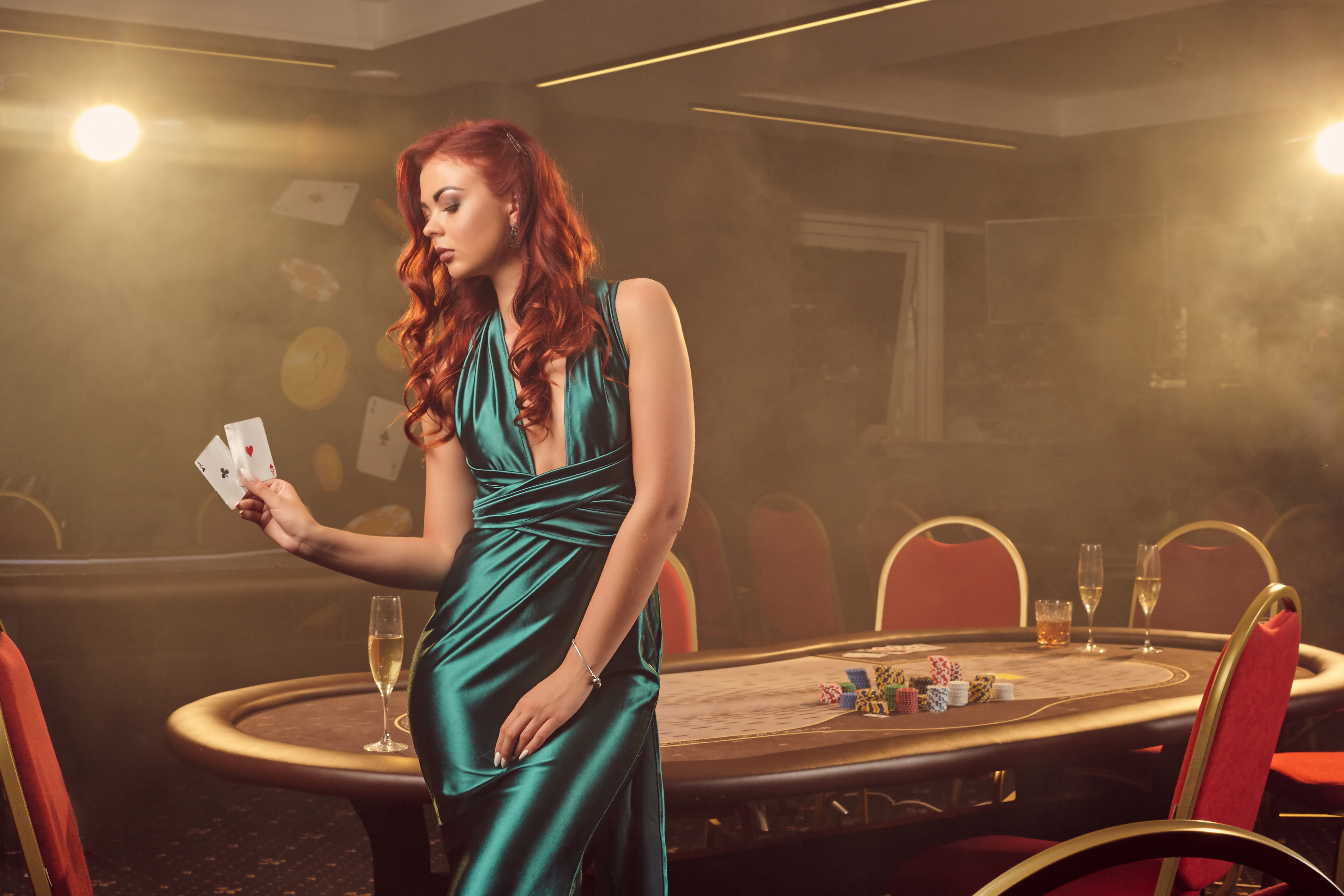 Attractive redheaded girl in a long blue satin dress is posing with two aces in her hand against a poker table in luxury casino. Passion, cards, chips, alcohol, win, gambling - it is as female entertainment. Smoke background.